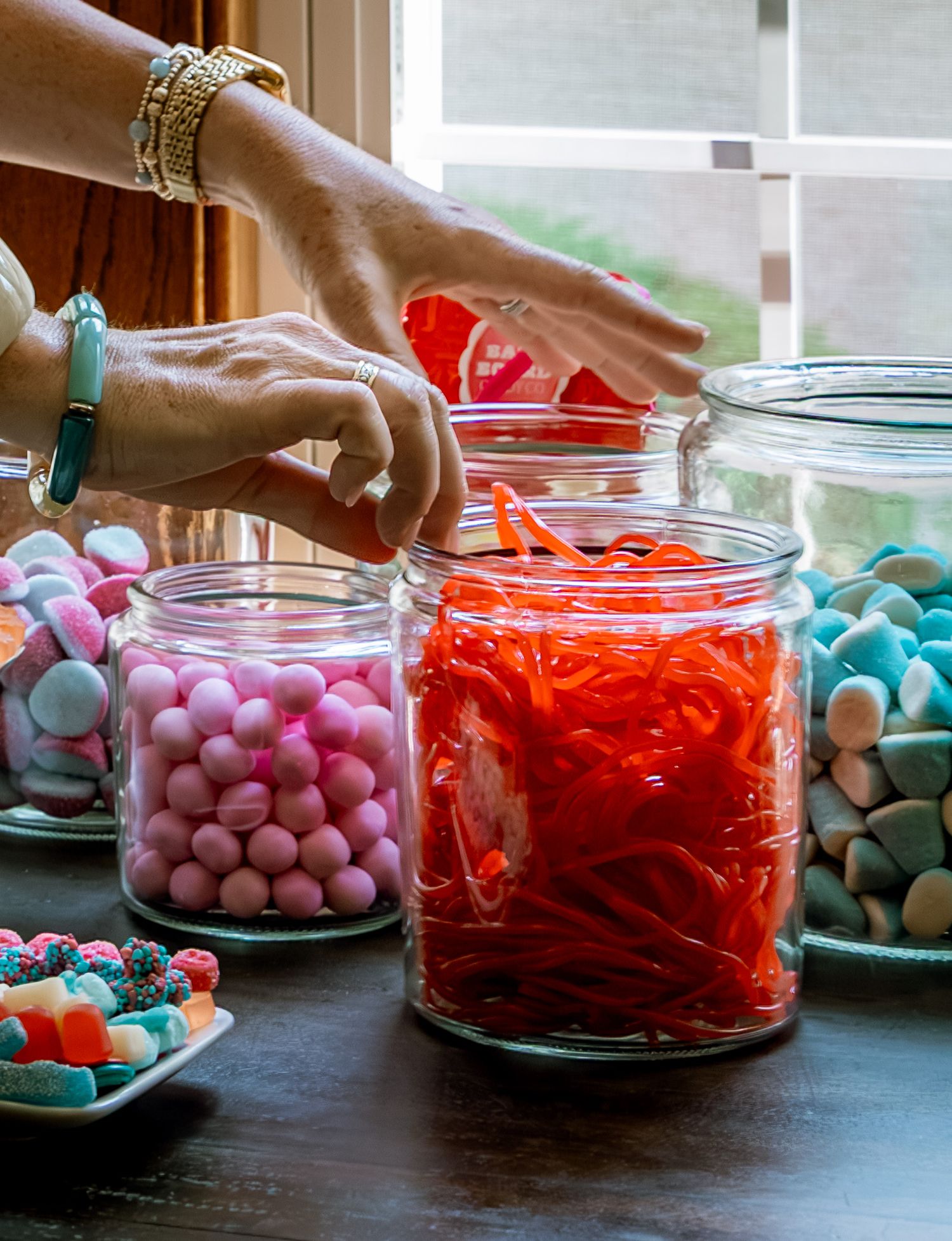Hands reaching into glass jars filled with colorful candies including red licorice, pink gumballs, and pastel marshmallows on a table.
