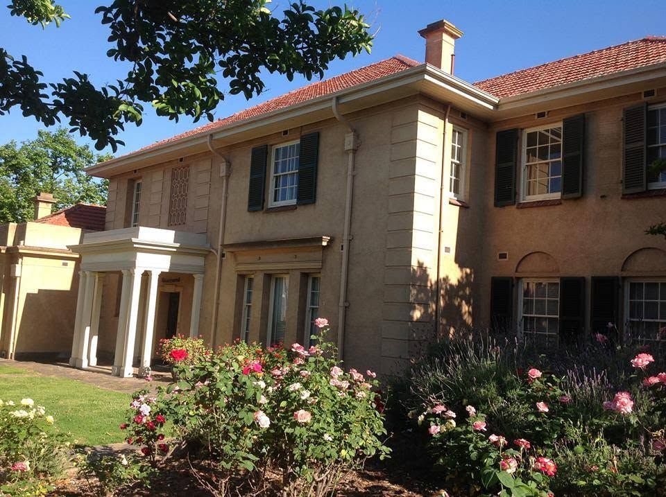 Historic two-story colonial mansion with rose garden, terracotta roof, and lush green trees on a sunny day.