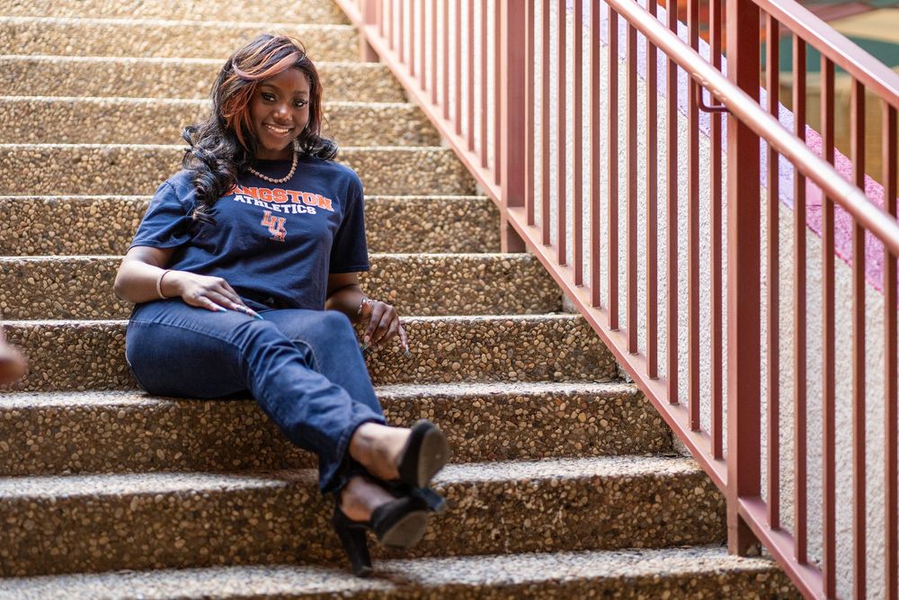 Smiling young woman in Langston University shirt sits relaxed on outdoor stadium steps.