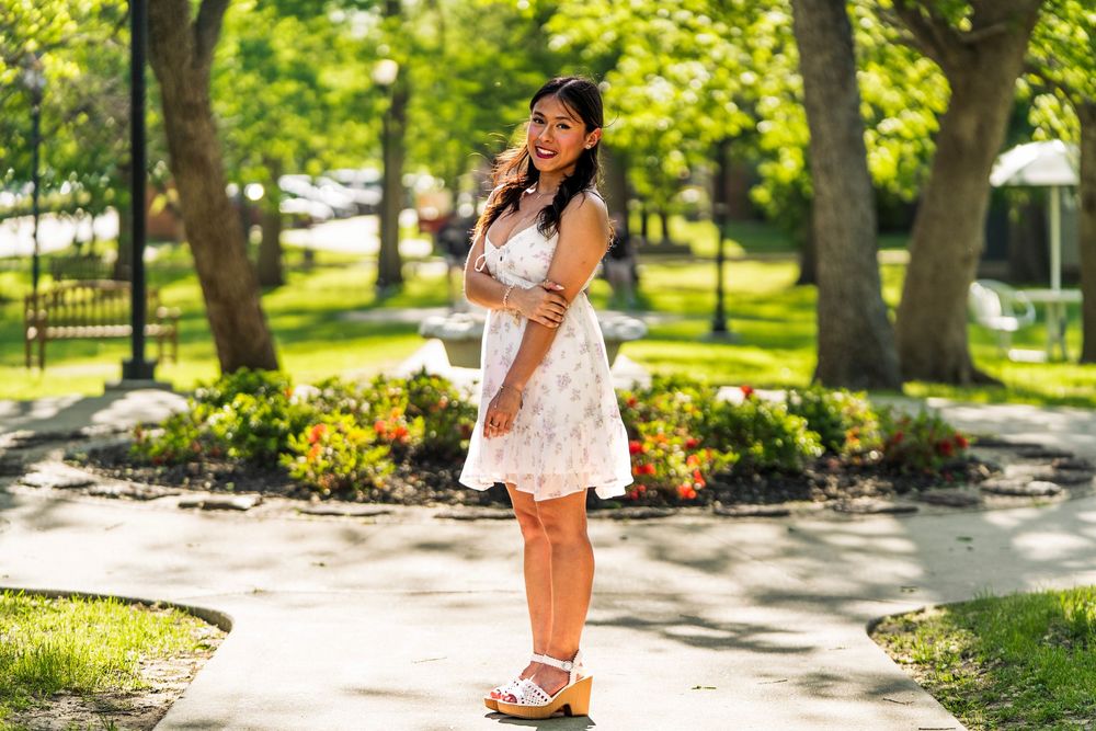 Young woman in floral white dress smiles while standing on sunny park pathway.