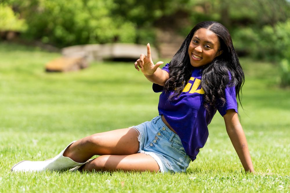 Smiling young woman in purple jersey sits on grass giving school sign outdoors.