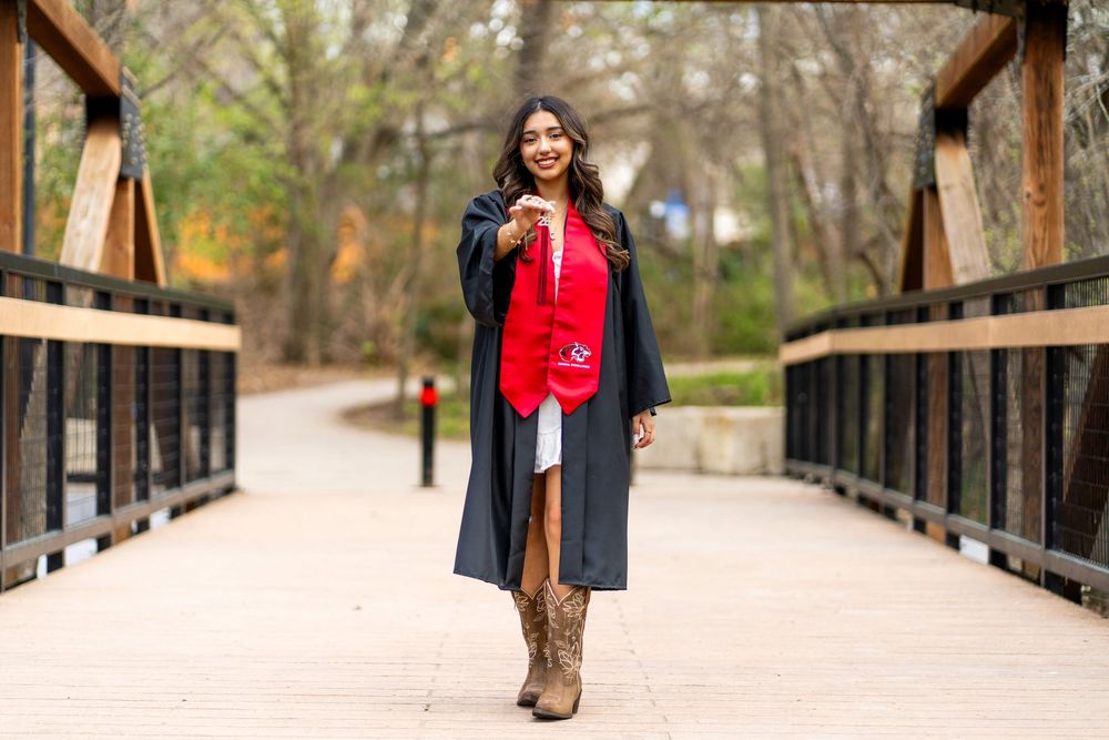 Graduate in black gown and red stole poses proudly on wooden bridge outdoors.