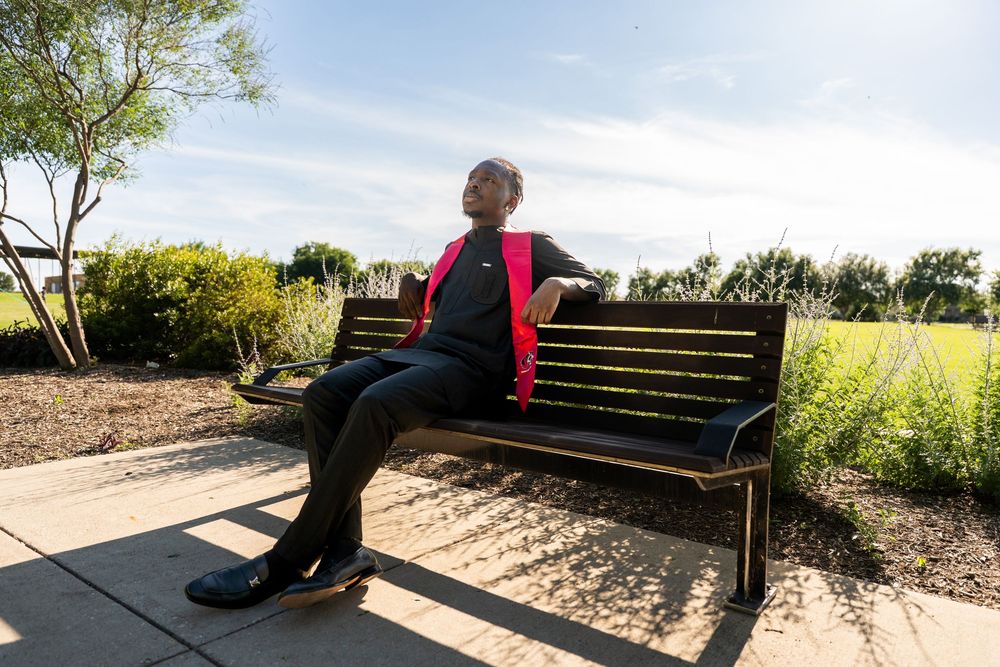 Young man in graduation attire relaxes on park bench under bright blue sky.