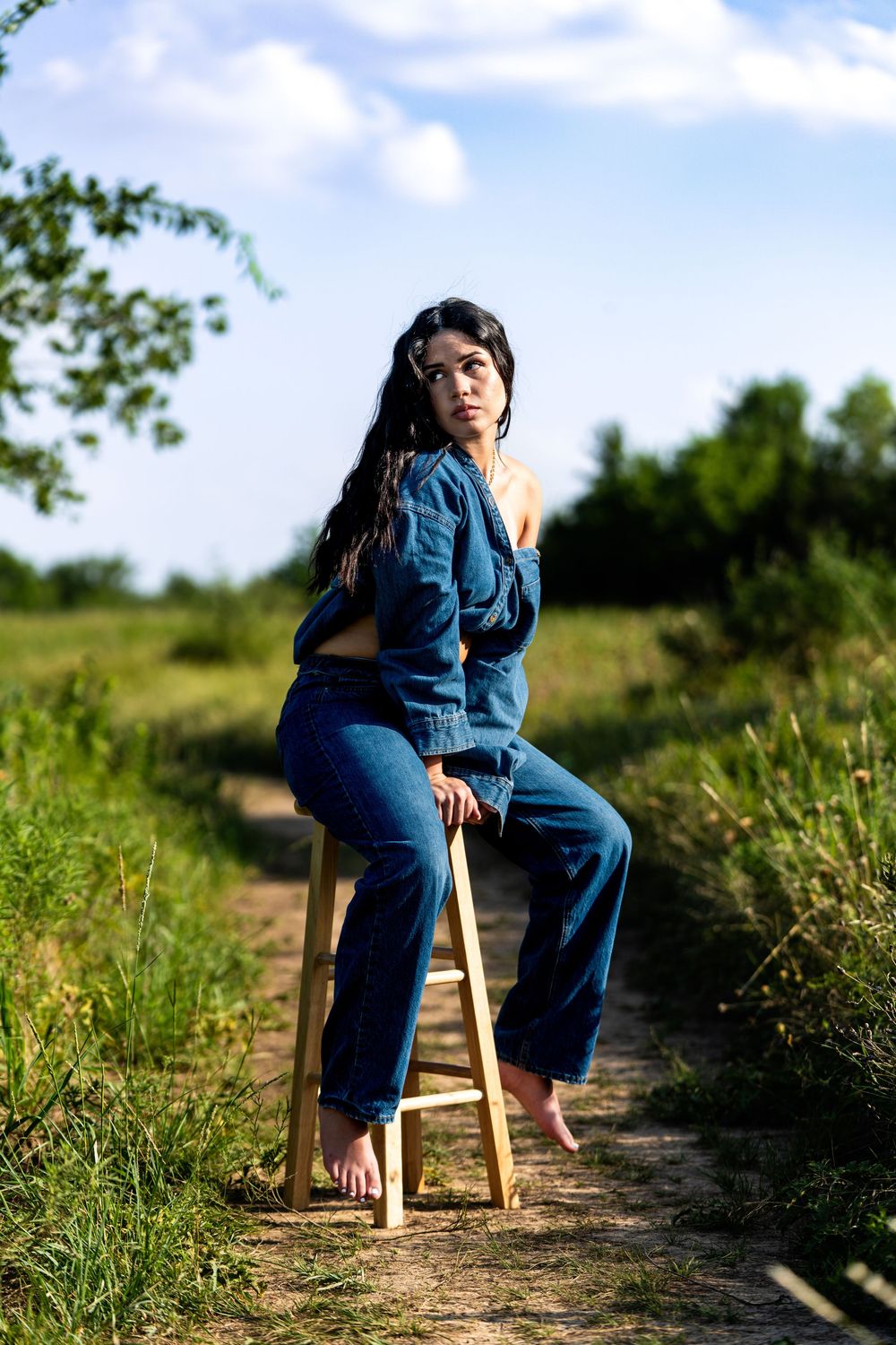 Young woman in denim outfit sits on stool on rural dirt path.