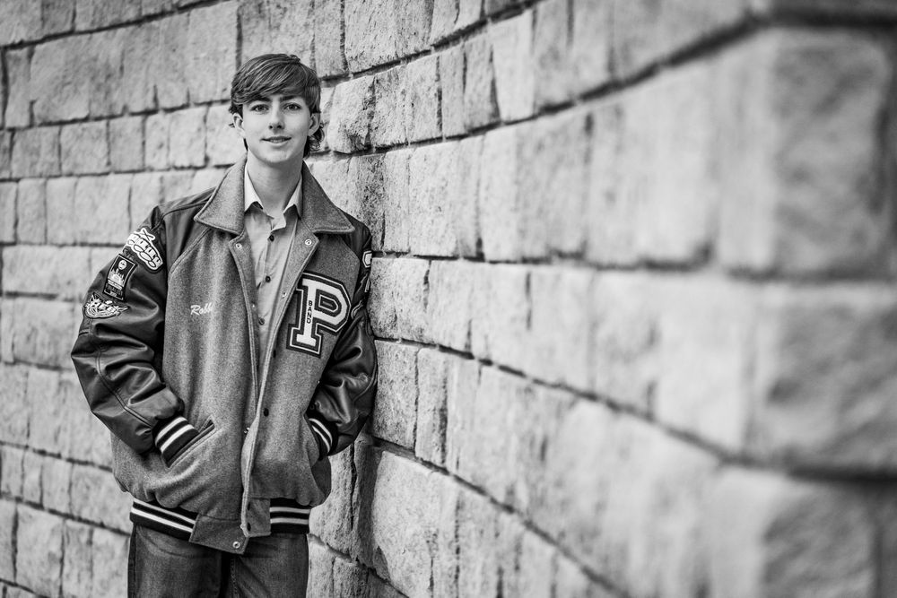Teen in varsity jacket leans against brick wall in black and white.