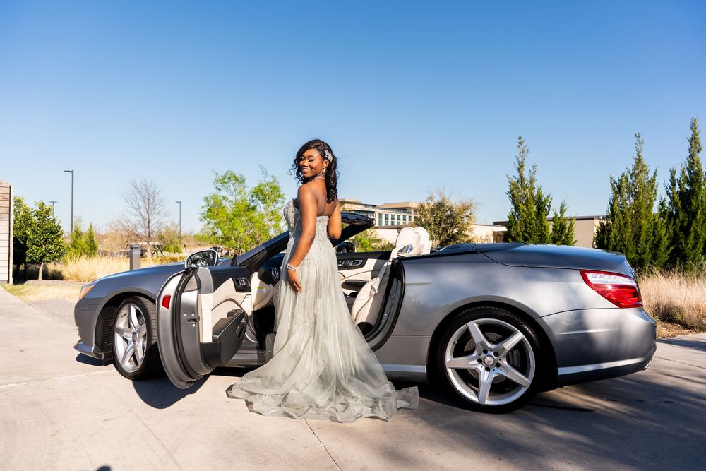 Young woman in elegant gown poses beside silver Mercedes convertible.