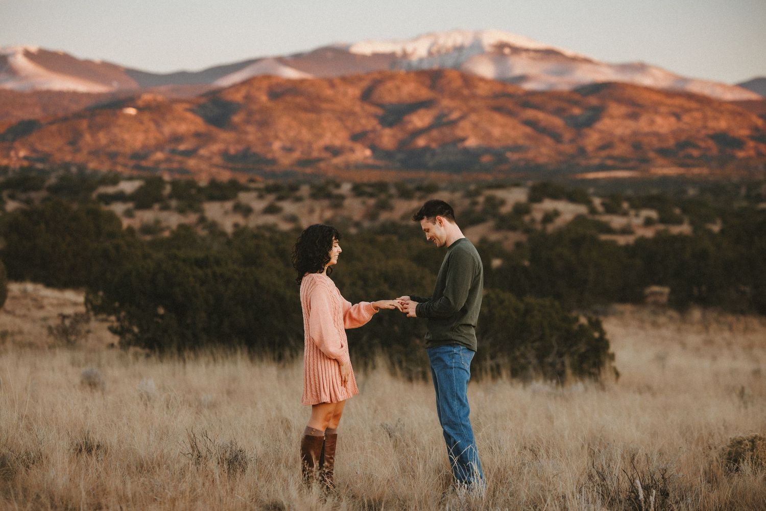 Couple sharing a romantic proposal moment in a golden field with snow-capped mountains at sunset.