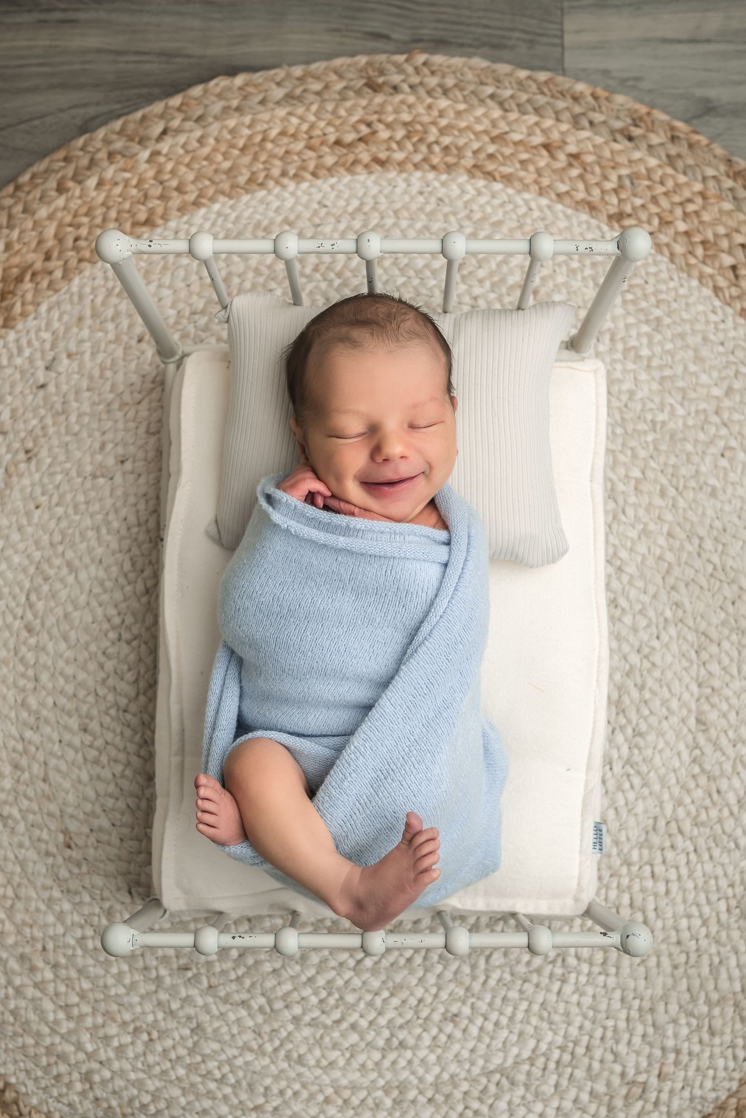 Smiling newborn baby swaddled in a light blue blanket lying on a tiny white miniature bed on a round woven rug.