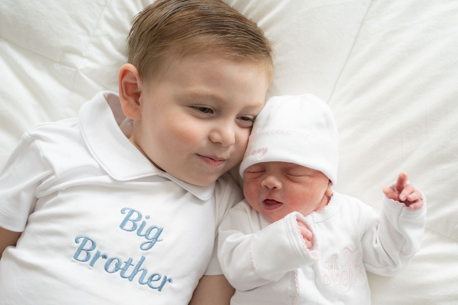 A toddler wearing a Big Brother shirt cuddles with a newborn baby in white, both lying on a white bed.