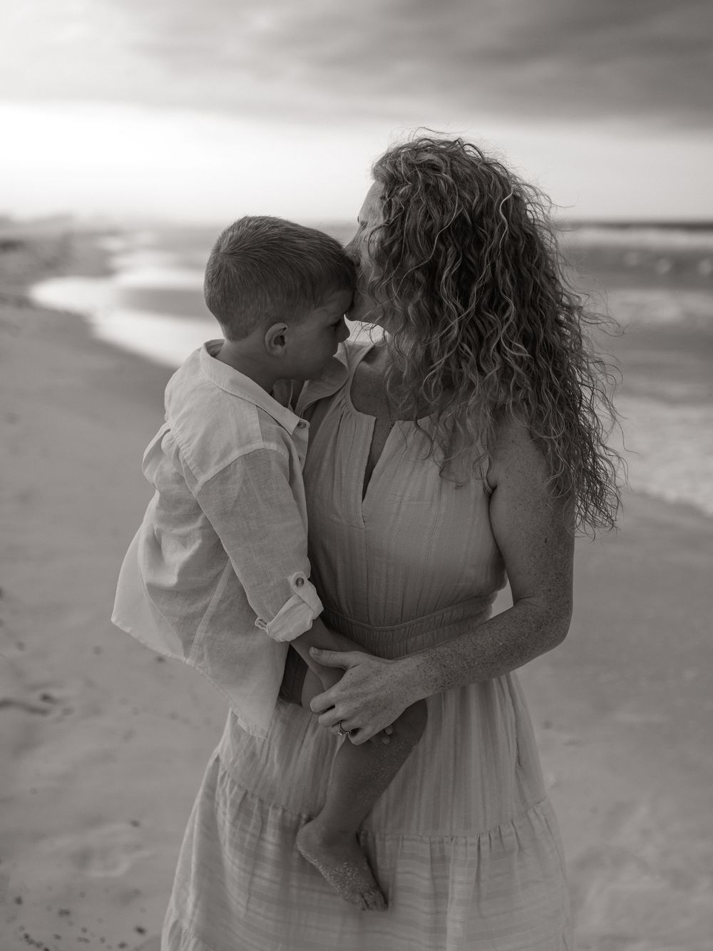 Black and white photo of a curly-haired woman holding a young boy on a beach at sunset.