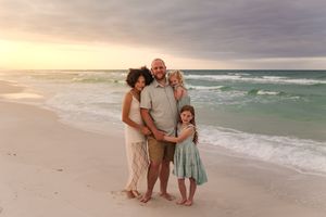 Family of four posing on a sandy beach at sunset with turquoise waves and pastel sky behind them.