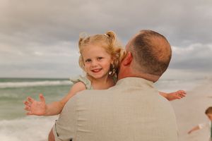 A smiling toddler girl with pigtails being held by a man on a sandy beach with ocean waves in the background.