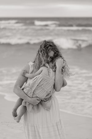 Black and white photo of a curly-haired mother holding her young child on a beach with ocean waves.