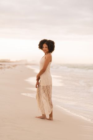 A woman in a flowing white lace dress stands barefoot on a sandy beach at sunset, smiling over her shoulder.