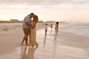 Couple kissing on a sandy beach at sunset while children play in the ocean waves behind them.