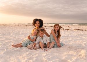 A mother and three children smile together on a white sandy beach at sunset in matching sage green outfits.