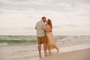 Couple embracing on a sandy beach with ocean waves, woman in gold dress and man in khaki shorts.