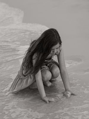 Black and white photo of a woman crouching on a sandy beach as ocean waves wash around her feet.