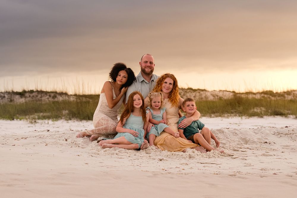 Family of six poses together on a white sand beach at sunset with sea oats and dramatic sky behind them.