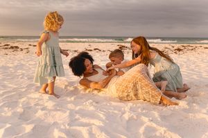 Family playing together on white sand beach at golden hour sunset, with children and mothers laughing in casual dresses.
