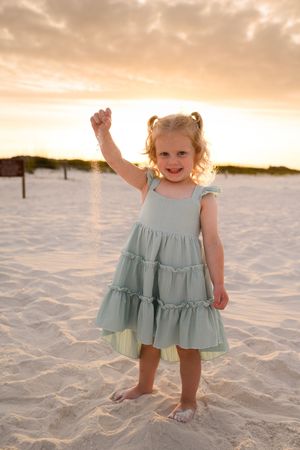 Young girl in mint tiered dress waves hand on sandy beach at golden sunset.