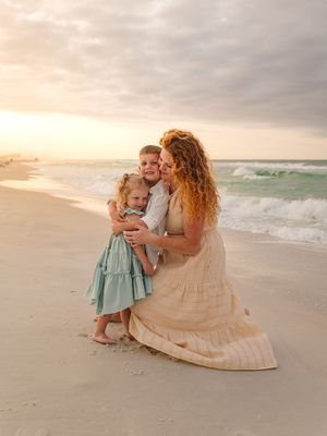 A mother with curly red hair hugs two young children on a sandy beach at golden sunset hour.