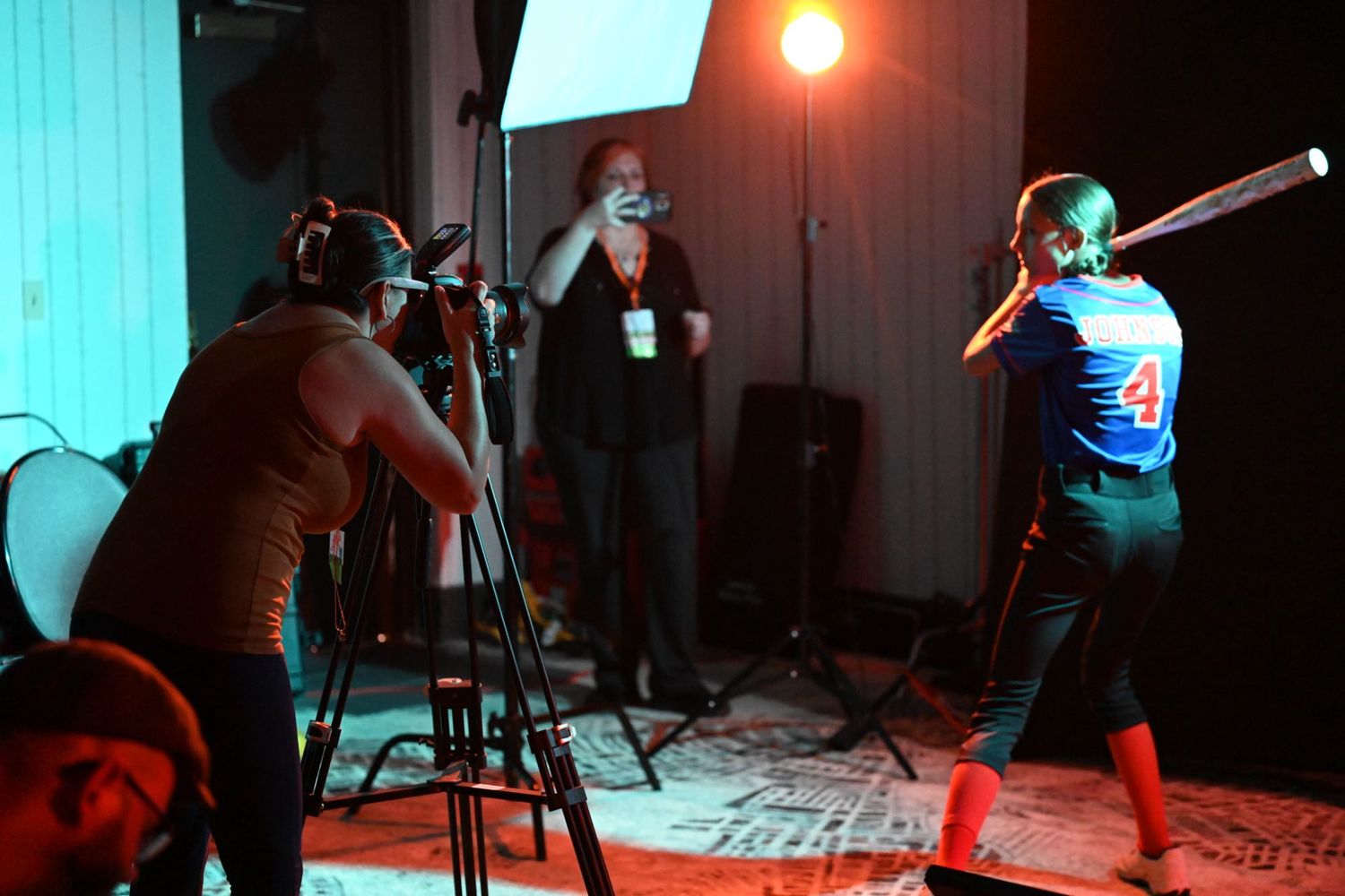 Photographer filming a baseball player in batting stance during an indoor photo shoot with studio lighting.