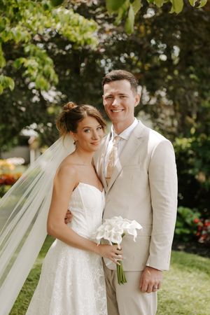 Bride in strapless lace gown with veil holds calla lily bouquet beside groom in beige suit outdoors.