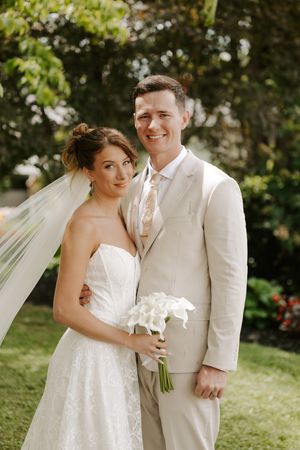 Bride and groom pose outdoors in garden, she holds white calla lily bouquet wearing lace gown with veil, he wears beige suit.