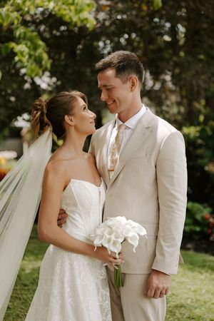 Bride and groom gazing at each other outdoors, she holds white calla lilies, he wears a beige suit with gold tie.