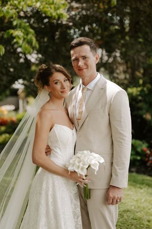 A bride in a white lace gown holding calla lilies stands with a groom in a beige suit outdoors.