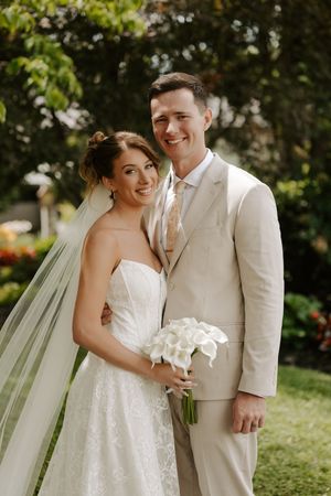 Smiling bride and groom pose outdoors, she holds white calla lily bouquet, wearing lace gown and veil, he wears beige suit.