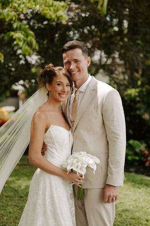 Bride and groom pose outdoors, she holds white calla lilies bouquet, wearing lace gown with veil, he wears beige suit.