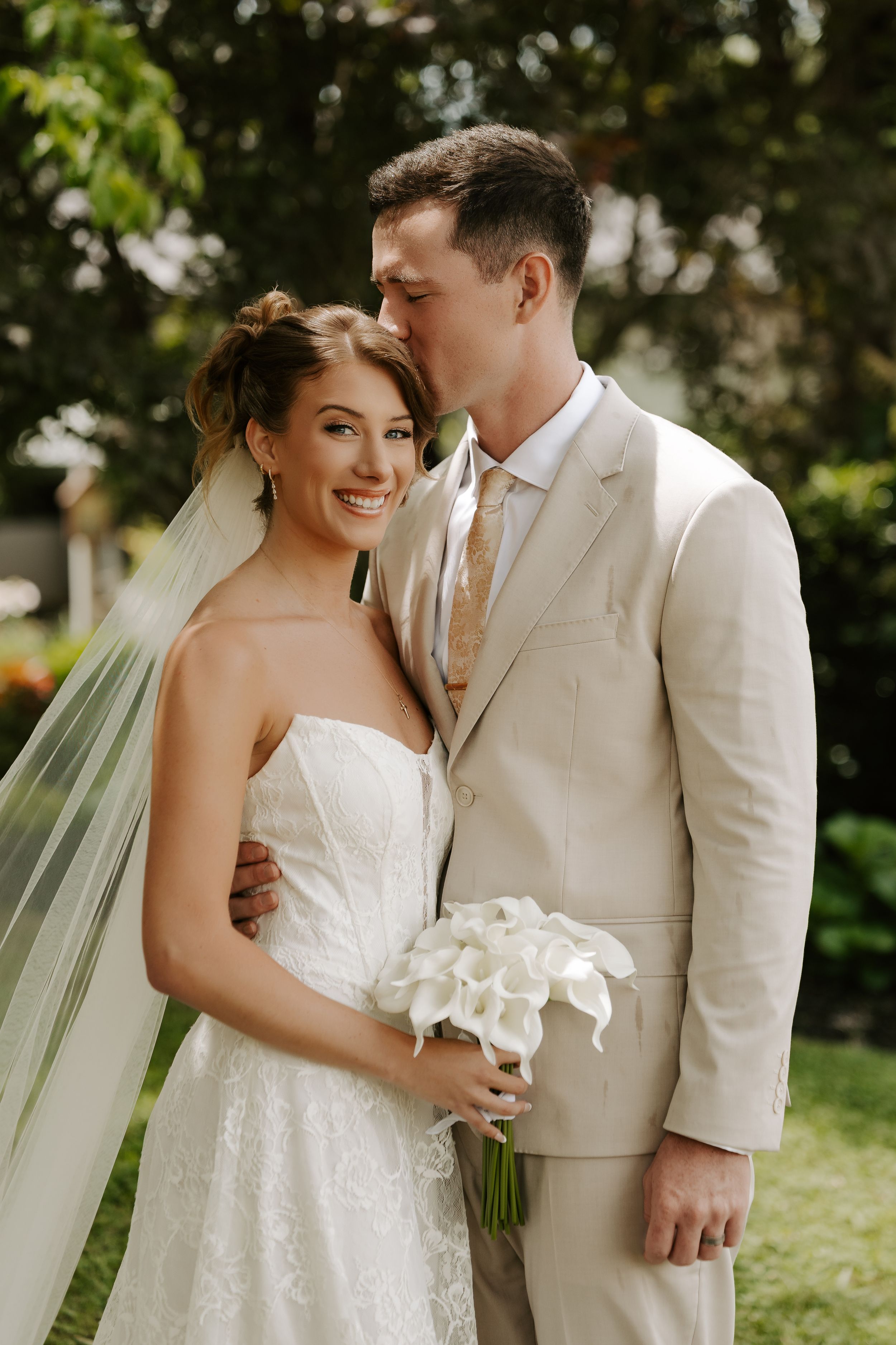Groom kissing bride on the head outdoors, bride smiles holding white calla lily bouquet in lace strapless gown.