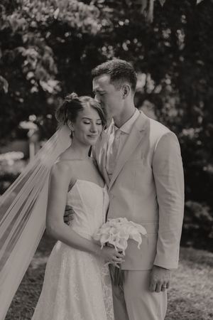Black and white photo of bride and groom embracing outdoors, bride holding white bouquet wearing lace gown with veil.
