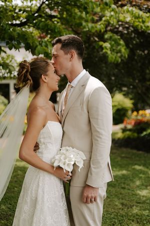 Bride and groom sharing a tender kiss outdoors in a lush garden, she in lace gown with veil, he in beige suit.