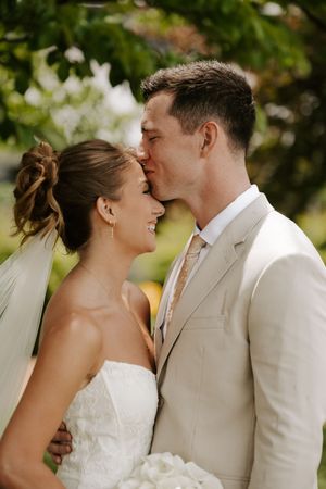 Groom in beige suit kissing smiling bride on forehead outdoors on wedding day.