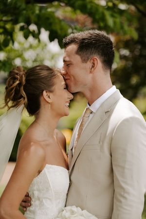 Happy bride smiling as groom in tan suit kisses her forehead during wedding portraits.