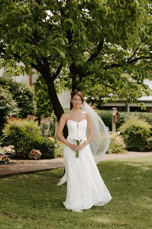 Bride in strapless white lace gown holding bouquet, standing on green lawn under large tree in garden.