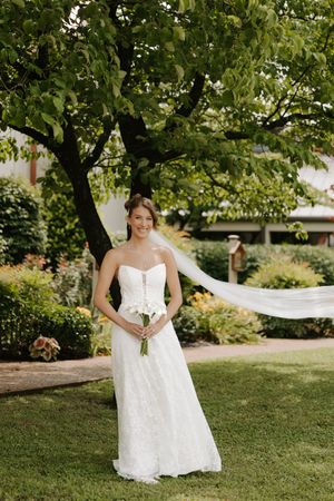 Smiling bride in strapless lace wedding gown holding white bouquet with veil flowing in garden.