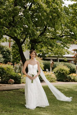 Smiling bride in strapless lace wedding dress holds white bouquet with veil trailing in garden.