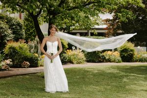 Bride in white gown with flowing veil caught by wind, holding bouquet, standing on green lawn with lush garden trees.