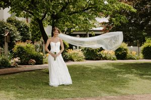 Bride in white gown with flowing veil caught in wind, standing on green lawn surrounded by lush garden trees.