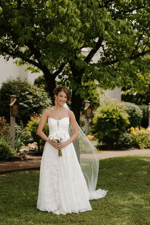 Elegant bride in strapless lace gown and flowing veil holds white bouquet in lush garden setting.