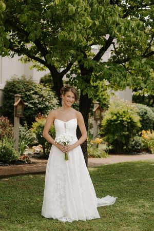 Bride in strapless lace wedding gown holding white bouquet standing in lush garden.