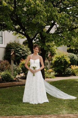 Elegant bride in strapless lace A-line wedding gown holding white bouquet, standing on green lawn beneath a tree.