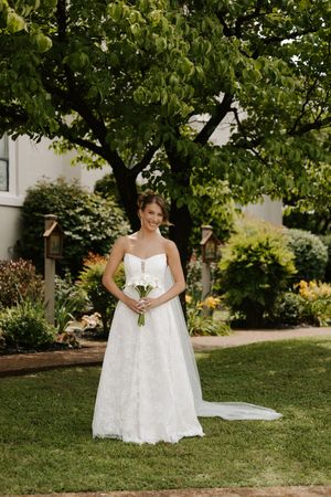 Bride in strapless lace wedding gown holds white bouquet while standing on green lawn beneath large tree in garden setting.