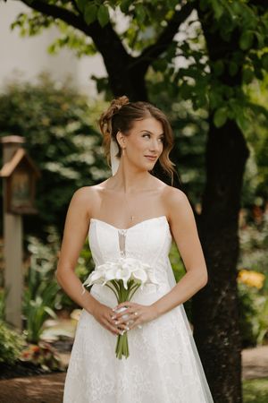 Elegant bride in strapless lace wedding dress holding white calla lily bouquet in lush garden setting.