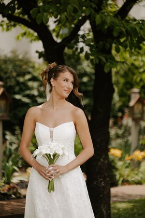 Elegant bride in strapless lace gown gazes sideways holding white calla lily bouquet in lush garden with trees behind her.