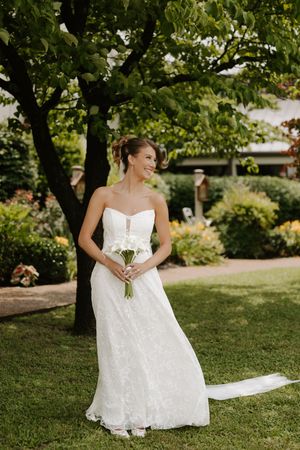 Bride in strapless lace wedding gown holding white bouquet, smiling in lush garden setting with green trees.